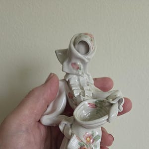 Op de afbeelding: Een wit porseleinen beeldje van een persoon met een hoofddoek en een blouse met ruches, die een klein kommetje vasthoudt. Het stuk heeft delicate roze bloemmotieven op de hoofddoek, het lijfje en de kom, met een groen en roze geruit schort.
