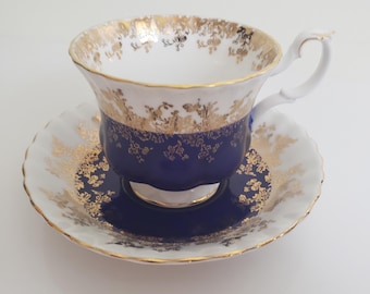 May include: A white teacup and saucer with a blue and gold floral pattern. The cup and saucer have a gold rim and a scalloped edge.