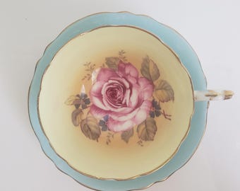 May include: A vintage teacup and saucer set. The cup and saucer are light blue with a scalloped edge and gold trim. The cup interior is yellow with a large pink rose floral design. A classic tea set.