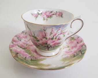 May include: A white teacup and saucer with a floral design. The cup and saucer feature a pink and green floral pattern with a gold trim. The design depicts a field of blooming trees.