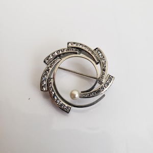 May include: A silver brooch with a circular design featuring a pearl in the center and a swirl of clear rhinestones around the edge.