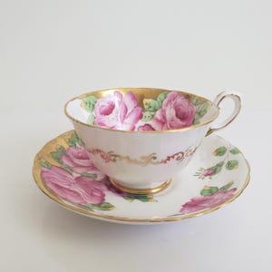 Op de afbeelding: Een witte theekop en schotel met roze rozenpatronen en gouden randen. De theekop heeft een gebogen oor en staat op een bijpassende schotel. Het servies is van porselein.