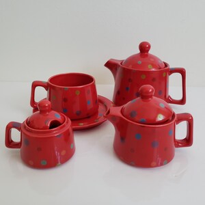 Peut inclure: Un service à thé rouge à pois avec une théière, un pot à crème, un sucrier, une tasse et une soucoupe. Les pois sont multicolores.