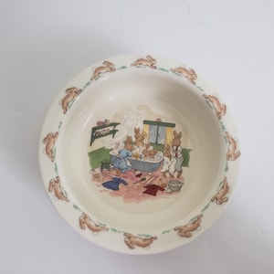 May include: A cream ceramic bowl with a rim decorated with brown bunnies. The bowl's interior features a charming illustration of bunnies bathing. The scene includes a bathtub, clothes, and other bath items. A green and pink border surrounds the illustration.