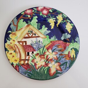May include: A blue and white ceramic plate with a raised design of a cottage with a water wheel, a bridge, and flowers. The plate has a blue background with green and yellow accents.