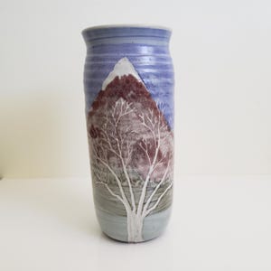 Puede incluir: Un jarrón de cerámica con un esmalte azul y blanco. El jarrón presenta una escena pintada de una montaña y un árbol con ramas blancas.