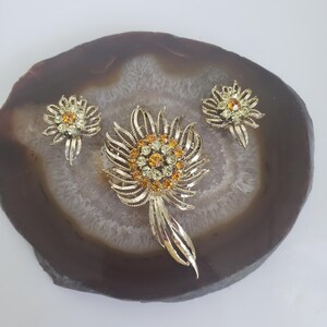 May include: A gold tone brooch and earrings set featuring a flower design with amber and clear rhinestones.