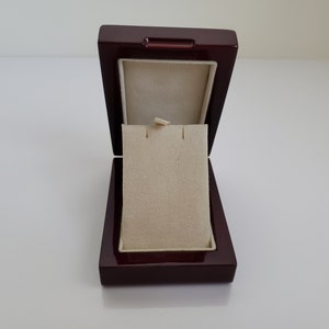 Puede incluir: Una caja de joyería de madera de color rojo oscuro con un interior de terciopelo beige. La caja está abierta y la tapa está levantada, revelando el forro de terciopelo.