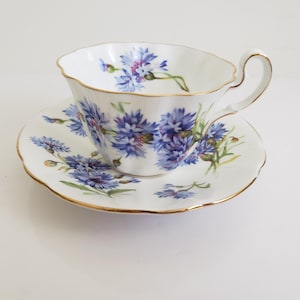 Op de afbeelding: Witte theekop en schotel set met een blauw bloemenpatroon. De theekop en schotel hebben een geschulpte rand en een gouden rand. Het bloemmotief toont blauwe en paarse bloemen met groene bladeren. Een klassieke theeset.