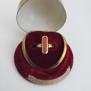 May include: A gold ring with a rectangular red stone set in the center. The ring is displayed in a heart-shaped velvet box.