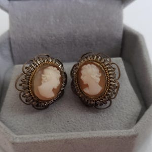 May include: A pair of vintage cameo earrings. Each earring features a profile of a woman carved in white against a peach-coloured background, set within an oval frame. The frames are surrounded by a decorative filigree design, with a gold-tone inner border.