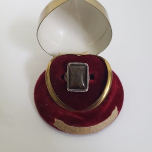 May include: A rectangular gemstone ring with a silver setting, presented in an open heart-shaped box. The ring features a dark, translucent stone. The box is gold-coloured with a red velvet interior.