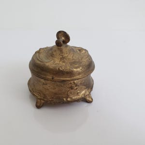 May include: A small, antique-style trinket box with a gold finish. The box has a rounded shape, a decorative lid with a small handle, and rests on three small feet. The surface has an ornate, textured design.