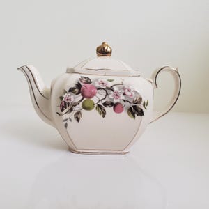 Peut inclure: Une théière blanche avec une bordure dorée et un motif floral représentant des pommes roses et vertes, des fleurs blanches et des feuilles vertes.