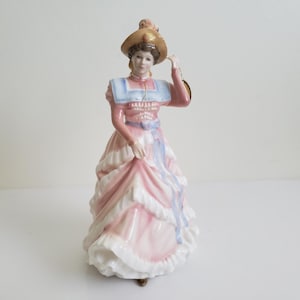 May include: A porcelain figurine of a woman in a long, layered pink and white dress, a blue shawl, and a tan hat. The figurine is holding the brim of her hat. The dress has a blue ribbon and the figurine is standing on a white surface.