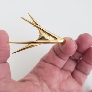 Könnte beinhalten: Eine goldfarbene abstrakte Brosche in Form eines Vogels im Flug. Die Brosche ist aus Metall und hat ein schlankes, modernes Design.