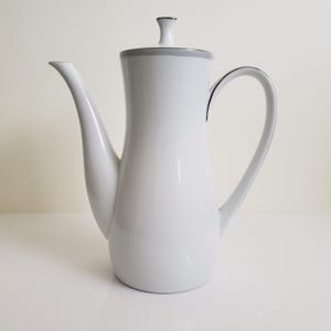 May include: A white ceramic coffee pot with a long spout and a curved handle. The pot has a grey rim and a matching lid with a small knob. The design is simple and elegant, suitable for serving coffee or tea.