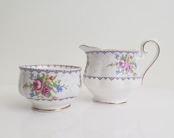 Può includere: Un set di zuccheriera e lattiera in porcellana bianca con un motivo floreale. Il set presenta un delicato disegno floreale con fiori rosa, blu e verdi su uno sfondo bianco. La zuccheriera e la lattiera hanno un bordo dorato.