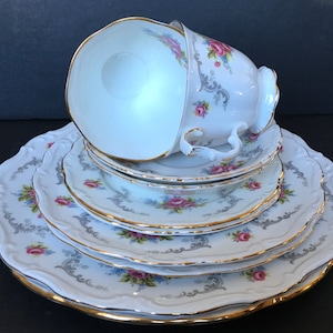 Puede incluir: Un juego de cuatro platos de porcelana blanca con un patrón floral y ribete dorado. Los platos están apilados uno encima del otro, con una taza de té y un platillo en la parte superior.