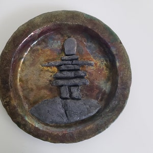 May include: A round ceramic plate with a raised, grey, stone-like sculpture of an inukshuk in the centre. The plate has a mottled, brown, green, and gold glaze.