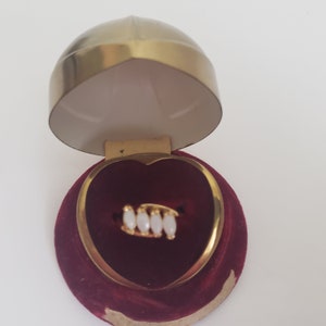 May include: A gold ring with white oval-shaped stones set in a heart-shaped velvet box with a gold lid.