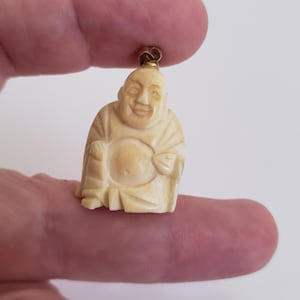 May include: A small, carved ivory figurine of a smiling Buddha wearing a robe. The figurine has a gold-coloured loop at the top for hanging.