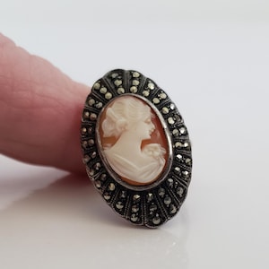 May include: Oval antique cameo ring. The ring features a cream-coloured shell carving of a woman's profile set against an orange background. The silver-toned setting is embellished with small, dark, sparkling accents.