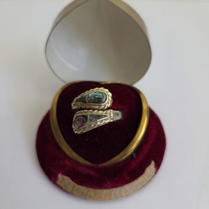 May include: A bypass ring featuring two teardrop-shaped inlays of iridescent abalone shell, each framed by a twisted metal border. The ring is displayed inside a heart-shaped jewellery box with a deep red velvet interior and a gold-toned metal rim.