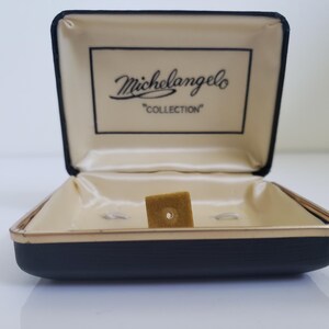 Puede incluir: Una caja negra abierta con un interior beige. La caja tiene un borde dorado y las palabras "Michelangelo COLLECTION" impresas en el interior de la tapa. Un pequeño trozo de fieltro marrón cuadrado se encuentra dentro de la caja.