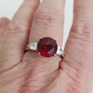 May include: A silver ring featuring a large, oval-shaped red gemstone, flanked by two smaller, pear-shaped, clear gemstones.