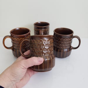 Puede incluir: Cuatro tazas de cerámica marrón con un patrón geométrico texturizado. Las tazas tienen un asa y una forma cilíndrica. Las tazas son de color marrón cálido y parecen ser de estilo vintage o retro.