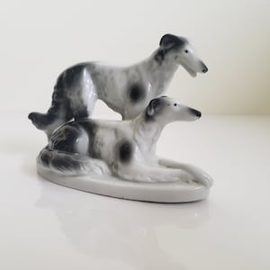 May include: A porcelain figurine of two greyhound dogs. One dog is standing on its hind legs, while the other is lying down. Both dogs are painted in black and white.