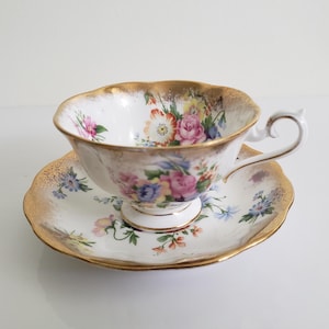 Puede incluir: Juego de taza y platillo de té blanco con diseño floral. La taza y el platillo tienen un borde dorado. La taza presenta un asa y un borde festoneado. El diseño floral incluye flores rosas, moradas y amarillas.