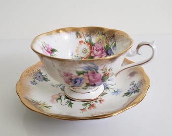 May include: A white teacup and saucer set with a floral design. The teacup and saucer have a gold trim. The teacup features a handle and a scalloped rim. The floral design includes pink, purple, and yellow flowers.