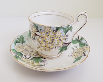 May include: White teacup and saucer set with a floral design. The teacup and saucer have a gold trim and a pattern of yellow flowers and green leaves. The teacup has a curved handle.