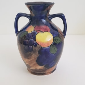 Puede incluir: Un jarrón de cerámica azul con dos asas y un diseño de frutas. El jarrón presenta una manzana amarilla, uvas rojas y moradas y un fondo marrón.