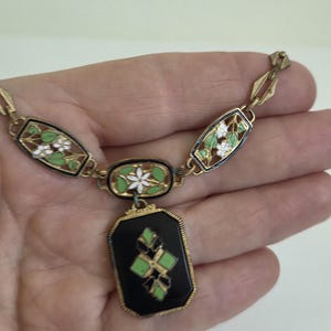 Puede incluir: Un collar de tono dorado con un colgante rectangular central negro con una incrustación geométrica verde y negra. La cadena consta de eslabones alternos, incluidos segmentos ovalados y rectangulares decorados con flores blancas y hojas verdes.