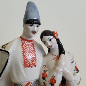 May include: A ceramic figurine of a couple in traditional dress. The man wears a pale blue hat and a white shirt with red geometric patterns. The woman has black hair and a white dress with red floral designs.