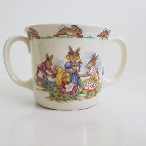 Puede incluir: Taza de cerámica color crema con dos asas, con una encantadora ilustración de conejos antropomórficos con vestidos y gorros. Los conejos se representan con cochecitos y sosteniendo conejitos bebés. Conejos marrones corriendo por el borde superior.