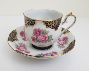 Puede incluir: Un juego de taza y platillo de té blanco con un diseño floral. La taza y el platillo tienen un borde negro y dorado con rosas rosadas y hojas verdes.