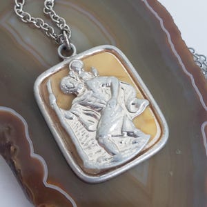 Può includere: Collana con pendente color argento. Il pendente rettangolare presenta un rilievo di una figura religiosa che trasporta un bambino, su sfondo giallo chiaro. Il pendente ha un bordo argentato e la catena è anch'essa argentata.