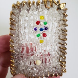Puede incluir: Un pequeño marco de vidrio transparente con un borde dorado. El marco está decorado con cuentas de vidrio de colores y una sola cuenta blanca en el centro.