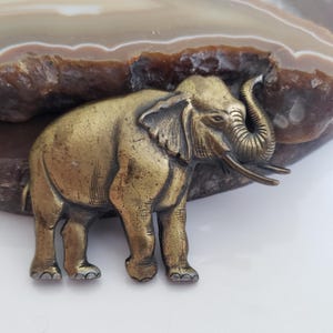 May include: A detailed brass elephant figurine with tusks and a raised trunk. The elephant is positioned in front of a brown and white stone. The figurine has a vintage aesthetic.