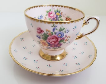 May include: A pink teacup and saucer set with a floral pattern and gold trim. The cup and saucer have a delicate, scalloped edge.