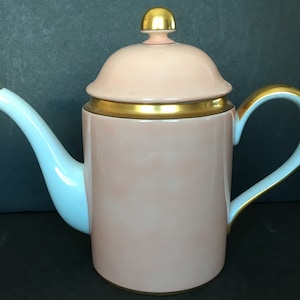 May include: A pink porcelain teapot with a gold trim and a gold knob on the lid. The teapot has a white spout and handle.