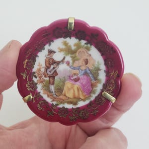 Puede incluir: Un pequeño plato redondo de porcelana rosa con un borde dorado y un diseño floral. El centro del plato presenta una escena pintada de una pareja en un entorno de jardín. El hombre está tocando la guitarra y la mujer sostiene una cesta de flores. El texto "Soyonar" es visible en la parte inferior del plato.