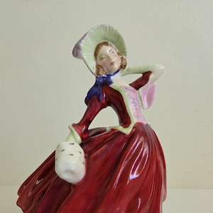 May include: A porcelain figurine of a person wearing a deep red dress with a pink bodice, a pale green bonnet with a dark blue ribbon, and holding a white muff with black spots. The figure has brown hair and is posed with one hand behind the head.
