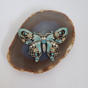 Puede incluir: Un broche de mariposa turquesa y dorado con detalles de cristal. El broche está sobre una rodaja de ágata marrón.