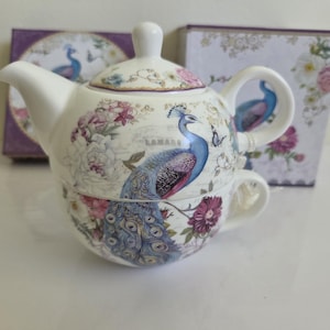 May include: A ceramic tea-for-one set featuring a teapot stacked on a teacup. The white porcelain is decorated with a vibrant blue peacock, pink and white floral patterns, and small butterflies. The set includes a matching lid and a box.
