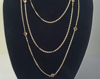 May include: A gold-toned, three-strand necklace with small, round, pinkish-brown gemstones. The necklace is displayed on a black mannequin, showcasing its layered design and delicate chain details. A stylish accessory for any outfit.